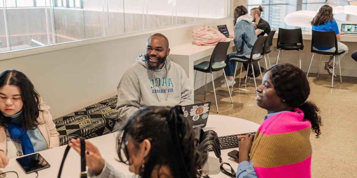 A group of students sits at  table talking. Some are working on computers.